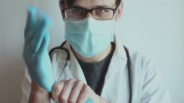 Gözlüklü ve tıbbi maskeli genç doktor hastaların muayenesine hazırlanırken steril eldiven takıyor. Genç bir doktor hasta muayenesi için hazırlanıyor. Koruyucu tulumlu bir doktor..