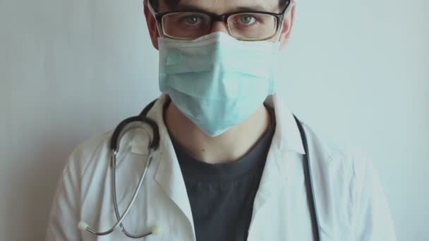眼鏡をかけた若いハンサムな医者は 彼の医療マスクを脱いで安心してため息をつく 若い医者は患者の回復を喜び 彼の保護マスクを脱いで安心してため息をつく — ストック動画