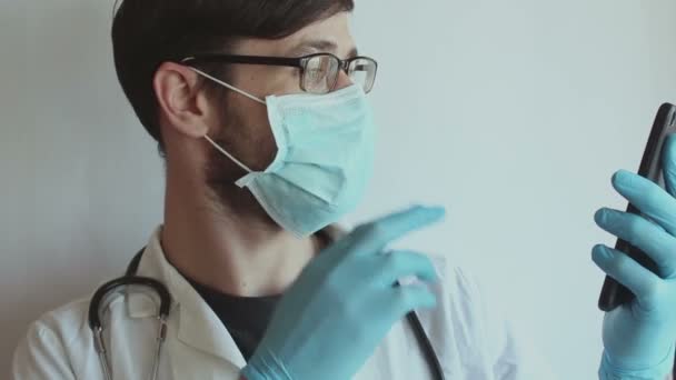 眼鏡をかけた若いハンサムな医者と医療面のマスクは 電話でビデオチャットの相談を患者に提供します 若い医師は 隔離中にビデオチャットを介して患者にオンライン相談を与える — ストック動画