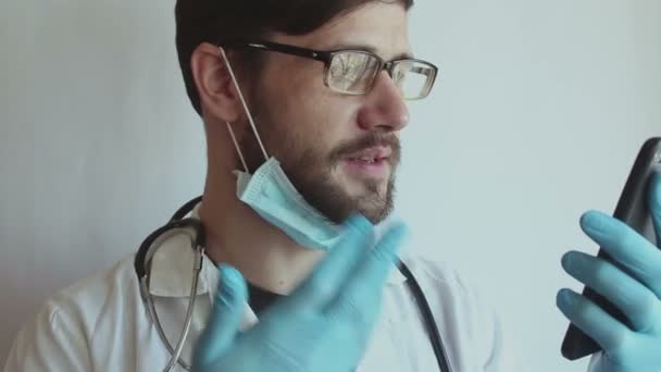 眼鏡をかけた若いハンサムな医者と医療面のマスクは 電話でビデオチャットの相談を患者に提供します 若い医師は 隔離中にビデオチャットを介して患者にオンライン相談を与える — ストック動画