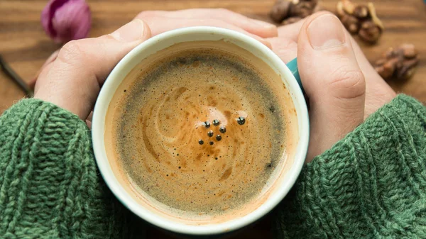 A men\'s hands are holding a cup of aromatic coffee. A cup of aromatic coffee stands on a wooden table. A man warms his hands with a cup of coffee.