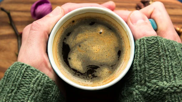 A men\'s hands are holding a cup of aromatic coffee. A cup of aromatic coffee stands on a wooden table. A man warms his hands with a cup of coffee.