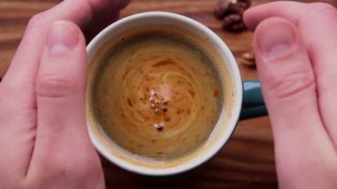 Bir erkek eli bir fincan aromatik kahve tutuyor. Ahşap bir masanın üzerinde bir fincan aromatik kahve duruyor. Bir adam bir fincan kahveyle ellerini ısıtır. Lezzetli süt köpüklü canlandırıcı kahve.