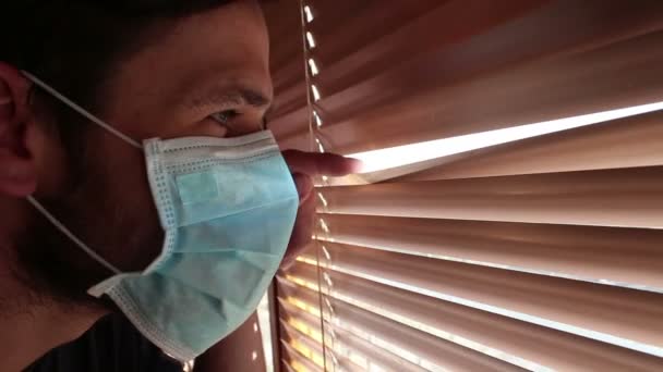 Un jeune beau mec avec un masque médical regarde à travers les stores. Un jeune homme dans un masque médical regarde par la fenêtre, étant sur l'isolement personnel. — Video