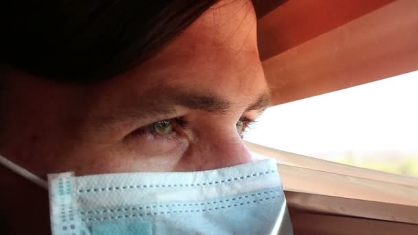 En ung stilig kille i ansiktsmask tittar genom fönsterpersiennerna. En ung kille i medicinsk mask tittar in genom fönstret och är isolerad.. — Stockvideo