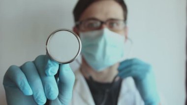  Gözlük takan ve tıbbi maske takan genç yakışıklı bir doktor hasta muayenesine hazırlanırken steteskop takıyor. Genç bir doktor steteskop tutuyor..