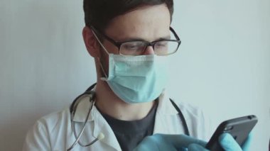 Gözlük takan ve tıbbi maske takan genç yakışıklı bir doktor hasta muayenesine hazırlanırken steteskop takıyor. Genç bir doktor steteskop tutuyor..