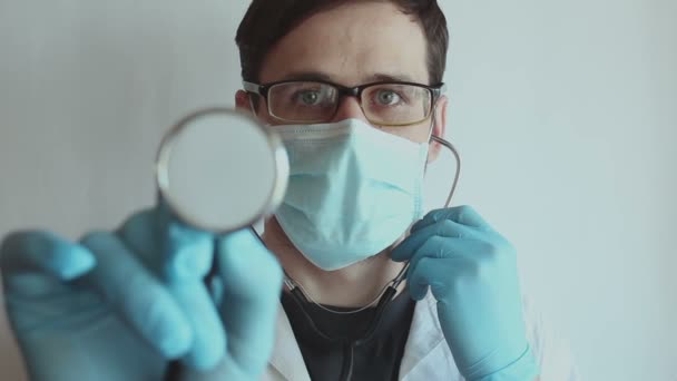 眼鏡をかけた若いハンサムな医者と医療面のマスクは 患者の検査の準備中に聴診器を装着します 若い医者が聴診器を持ってる — ストック動画