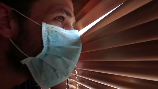 En ung stilig kille i ansiktsmask tittar genom fönsterpersiennerna. En ung kille i medicinsk mask tittar in genom fönstret och är isolerad.. — Stockvideo