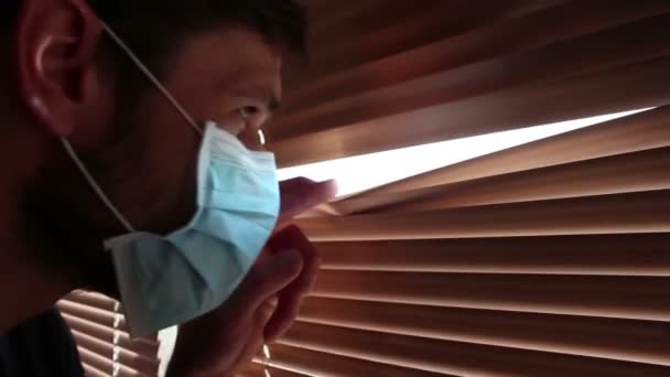 Een jonge knappe man met een medisch gezichtsmasker kijkt door de gordijnen. Een jongeman met een medisch masker kijkt door het raam, op zelfisolatie.. — Stockvideo