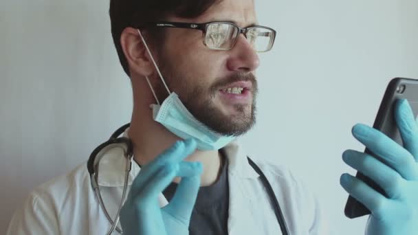 Un jeune beau médecin portant des lunettes et un masque médical fournit à un patient une consultation de chat vidéo au téléphone . — Video