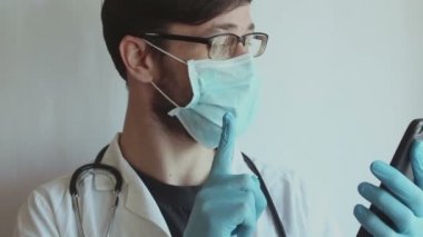 Gözlük takan ve tıbbi maske takan genç yakışıklı bir doktor telefonda bir hastaya görüntülü konuşma danışmanlığı sağlıyor..