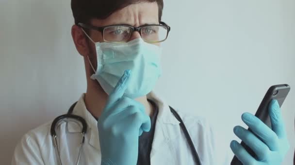 Ένας νεαρός όμορφος γιατρός που φοράει γυαλιά και μια ιατρική μάσκα προσώπου παρέχει σε έναν ασθενή μια συμβουλευτική βίντεο συνομιλίας από το τηλέφωνο. — Αρχείο Βίντεο