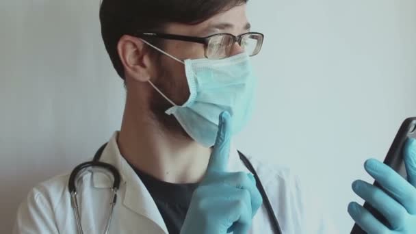 En ung stilig läkare bär glasögon och en medicinsk ansiktsmask ger en patient med en videochatt samråd via telefon. — Stockvideo