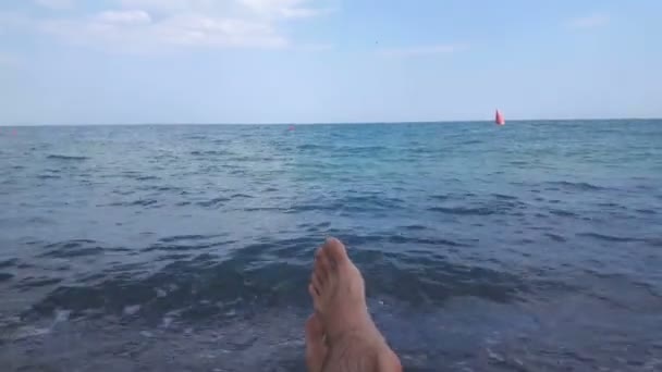 Vista de las piernas de un hombre acostado en la orilla del mar. Piernas masculinas sobre un fondo de mar azul.Un hombre disfruta de unas vacaciones en una playa de guijarros . — Vídeos de Stock