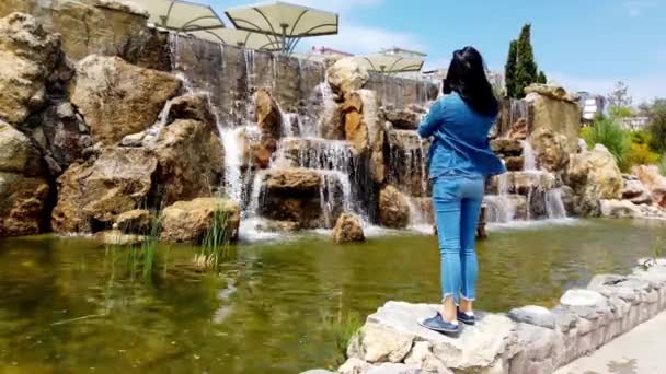 Junge Frau mit langen dunklen Haaren am Ufer des dekorativen Wasserfalls. — Stockvideo