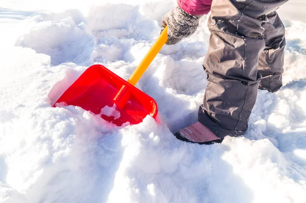 El niño cava una pala de nieve. Niño con las manos en manoplas sostiene una pala roja . — Foto de Stock