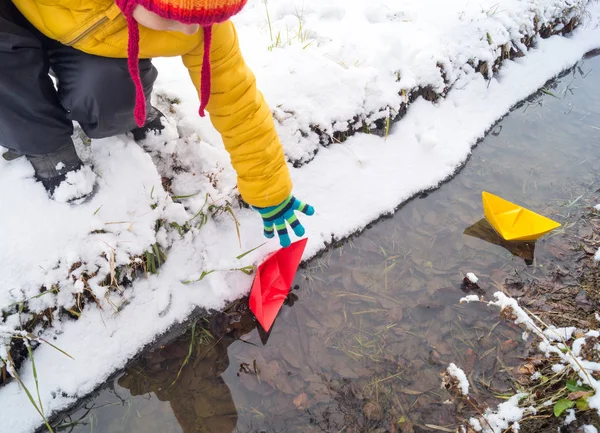 Niño busca un barco de papel a lo largo de un arroyo en invierno — Foto de Stock