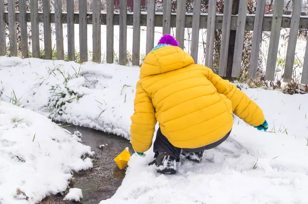 Niño busca un barco de papel a lo largo de un arroyo en invierno — Foto de Stock