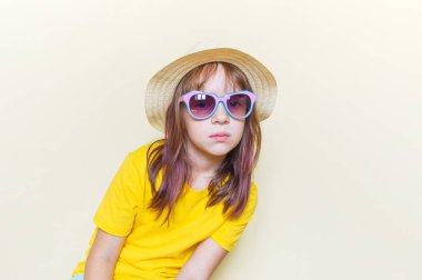 The child sits in the room on the floor emotion on his face. Little girl in a yellow T-shirt, glasses and a straw hat.
