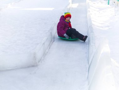 Çocuk buz kaydırağından yuvarlanıyor. Parlak elbiseler gökkuşağı şapkası.