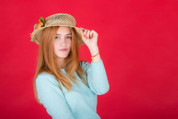 Portret pięknej, młodej rudej kobiety w słomkowym kapeluszu, dziewczyny pozującej na czerwonym tle w pracowni. — Zdjęcie stockowe