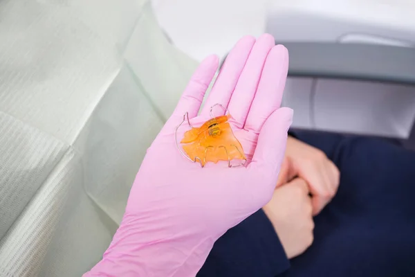 El dentista sostiene en su mano una placa de ortodoncia anatómica hecha de acrílico naranja y metal en forma de mandíbula. Primer plano — Foto de Stock