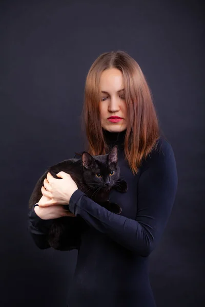 Rothaarige junge Frau in schwarzer Kleidung vor schwarzem Hintergrund mit schwarzer Katze — Stockfoto