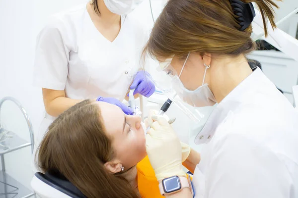 Un dentista en su consultorio o consultorio trata a una paciente con un asistente en máscaras y guantes — Foto de Stock