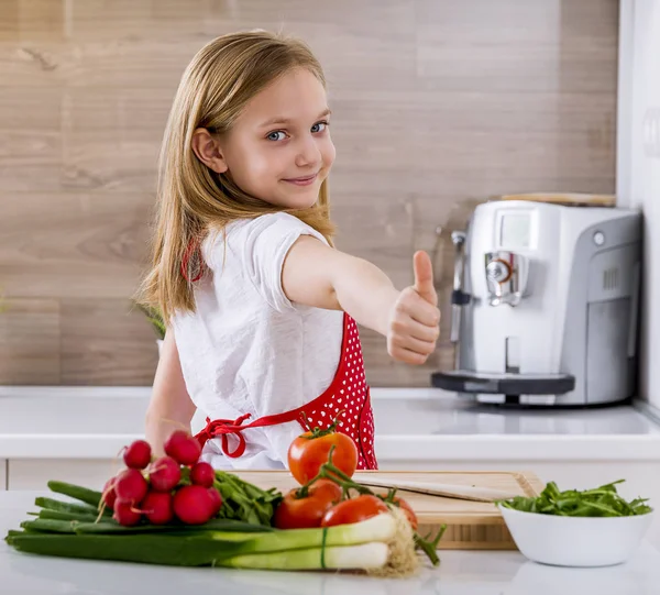 Menina bonita cozinhar na cozinha e mostrando polegares . — Fotografia de Stock