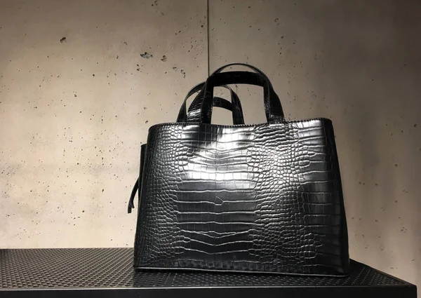Raftaki siyah deri taklit timsah çantası. Beton arka planda lüks bir mağazanın vitrininde iki tutacağı olan zarif bir kadın çantası. Moda kadın aksesuarları. Moda konsepti