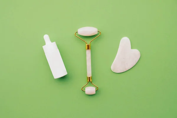 Jade stone roller made of pink quartz and cosmetic serum bottle for lymphatic drainage face massage. Top view on pastel green background, copy space, close up. Beauty and body care.