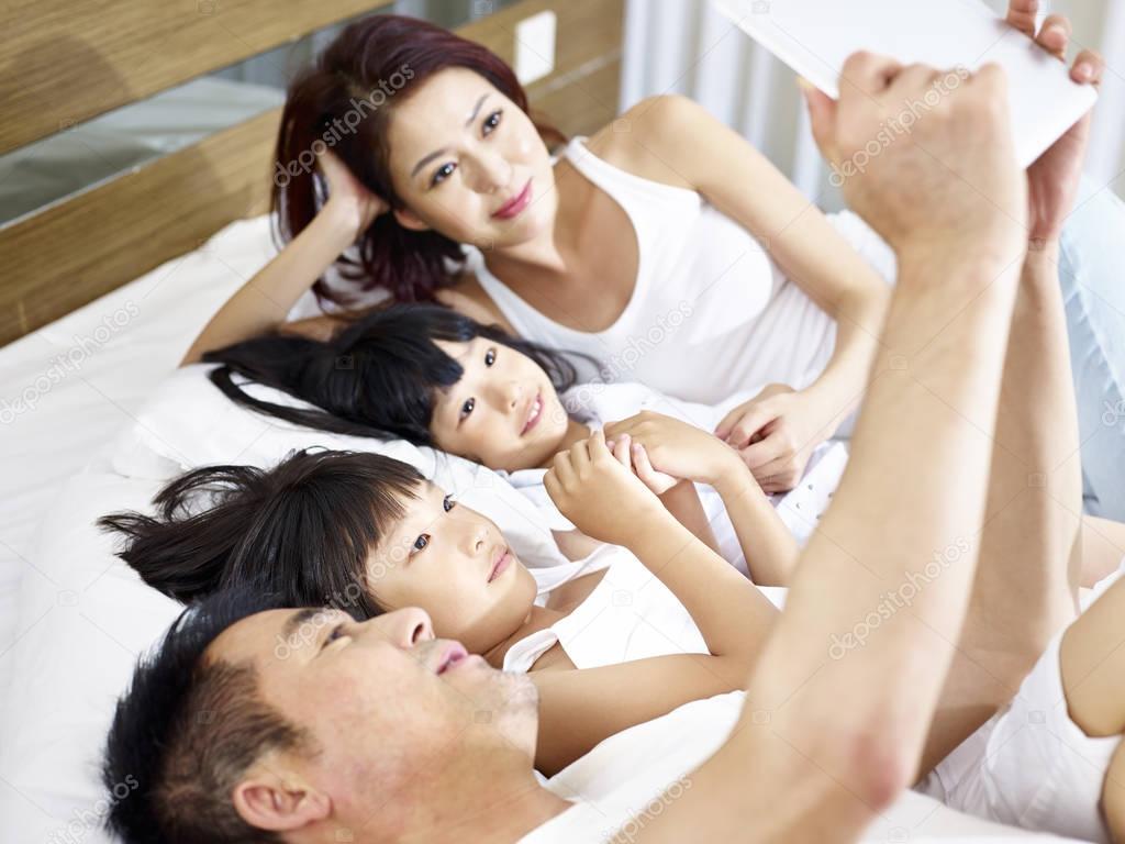 asian family with two children using digital tablet in bed