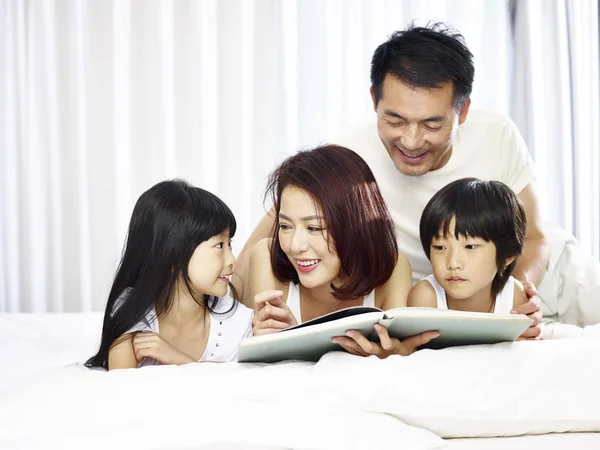 Asiática família com duas crianças lendo livro na cama — Fotografia de Stock