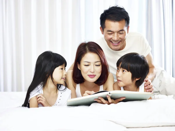 Asian couple and two children reading book together in bed — Stock Photo, Image