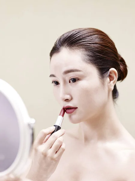 Young asian woman looking in the mirror while applying lipstick — Stock Photo, Image