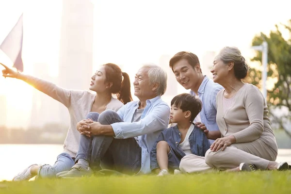 Üç kuşak mutlu Asyalı aile çimlerin üzerinde oturmuş parktaki alacakaranlıkta eğleniyorlar.