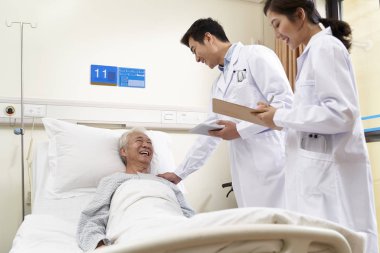 İki genç Asyalı doktor başucunda yaşlı bir adamla konuşuyor.