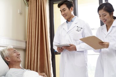 İki genç Asyalı doktor başucunda yaşlı bir adamla konuşuyor.