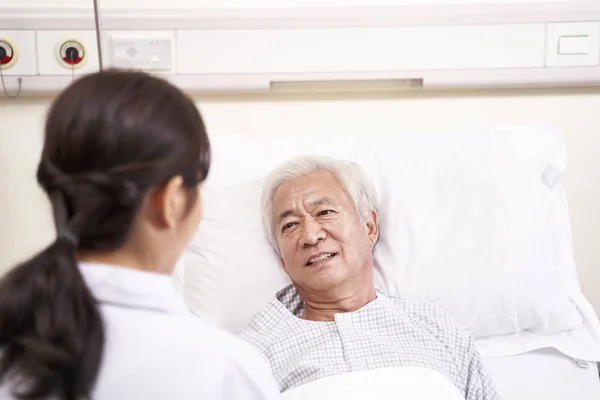 Азіатський старий лежить у ліжку, розмовляючи з молодою жінкою-лікарем у шлангах — стокове фото