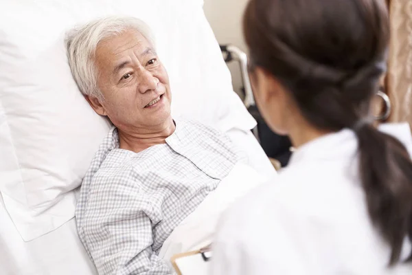 Asian old man lying in bed talking to young female doctor in hos — Stock Photo, Image