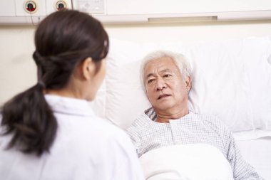 Genç Asyalı doktor başucundaki yaşlı hastayla konuşuyor.