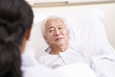 Genç Asyalı doktor başucundaki yaşlı hastayla konuşuyor.