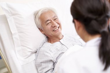 Genç Asyalı doktor başucundaki yaşlı hastayla konuşuyor.