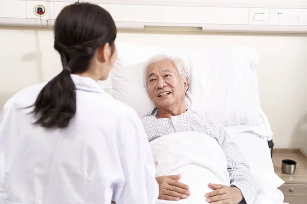 Asian old man lying in bed talking to young female doctor in hos — Stock Photo, Image