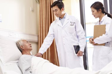 Kıdemli Asyalı erkek hasta yatağında uzanıp hastane koğuşundaki doktorlarla konuşuyor.