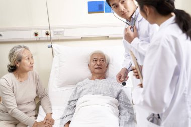 Genç Asyalı doktor yaşlı hastayı muayene ediyor. Hastane koğuşunda tıbbi durumunu tartışıyorlar.