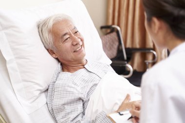 Genç Asyalı doktor konukevi koğuşunda yatağın yanında kıdemli bir hastayla konuşuyor.
