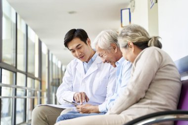 Genç Asyalı doktor hastane koridorunda dijital tablet kullanan yaşlı çift hastalarla test sonuçlarını ve teşhisi tartışıyor.