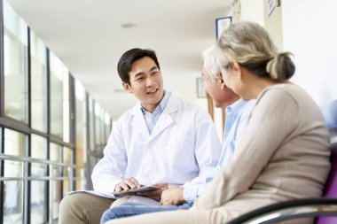 Dost canlısı genç Asyalı doktor hastane koridorunda yaşlı bir çiftle konuşuyor.